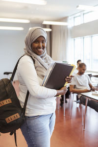 Portrait of smiling woman in hijab standing with books at university classroom
