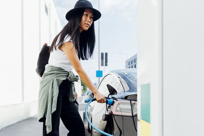 Young woman wearing hat charging electric car at vehicle station