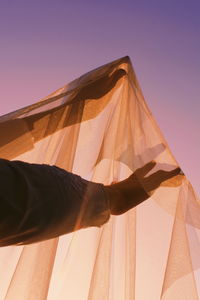 Cropped hand holding fabric against clear sky during sunset