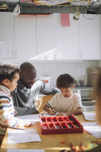 Male teacher assisting boy while writing on paper at bench in classroom