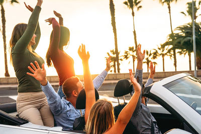 Friends with arms raised sitting in convertible against sky during sunset