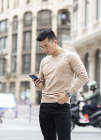 Masculine asian male standing with hand in pocket and browsing mobile phone in city while looking down