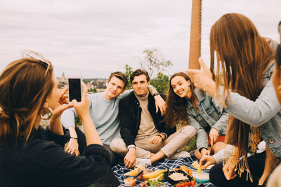 Woman photographing friends while enjoying picnic together against sky