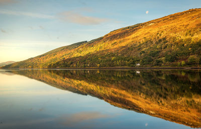 Scenic view of calm lake by hill against sky during sunset