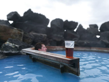 Man sitting on rock by swimming pool against sky