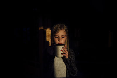 Girl with eyes closed holding mug while standing in darkroom at home