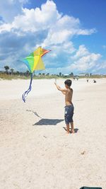 Full length of shirtless boy flying colorful kite at beach during summer