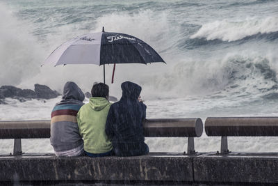 Rear view of friends sitting on retaining wall against sea during rain