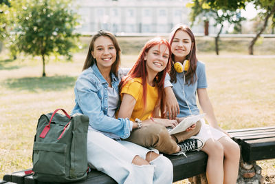 Three schoolgirls are sitting on a park bench, talking, preparing together with the lesson. 