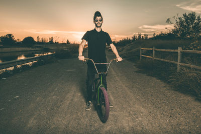 Portrait of young man riding bicycle while standing against sky during sunset