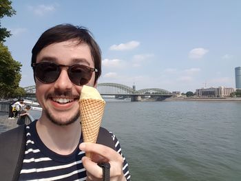 Portrait of young man holding ice cream against river against sky