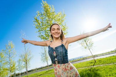 Portrait of young woman with arms raised standing against blue sky