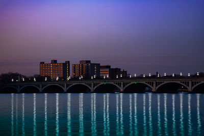 Bridge over river by illuminated buildings against sky at night