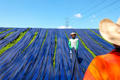 Man drying blue fabrics on grassy field against blue sky during sunny day