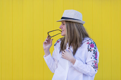 Woman holding cigarette while standing against yellow wall