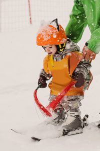 Cute attentive little kid in warm outerwear and protective helmet and vest holding brushes and balancing on snowy mountain slope while practicing skiing with cropped anonymous instructor