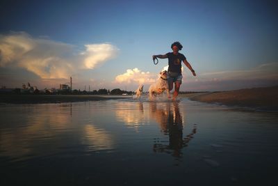 Woman running with dogs in lake against sky during sunset