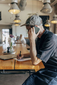 Male owner writing on notepad while talking on smart phone at cafe