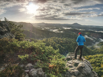 Photographer in blue sweatshirt working with mirror camera and tripod on peak of rock. dreamy fog