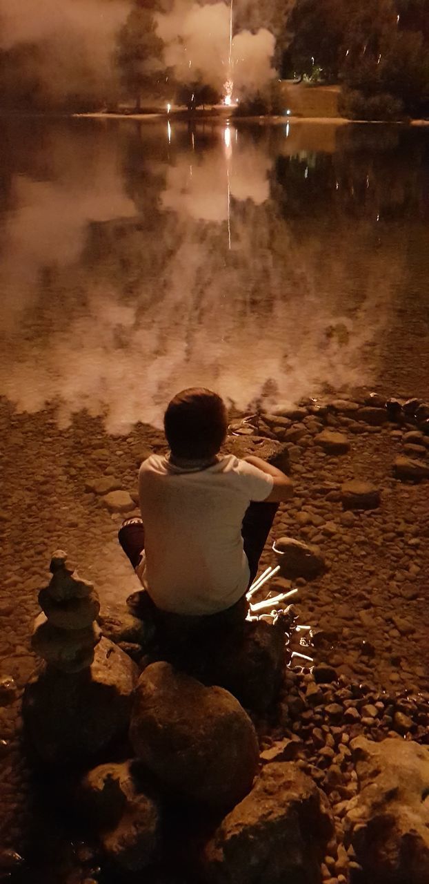 REAR VIEW OF BOY SITTING ON ROCK BY ILLUMINATED WATER