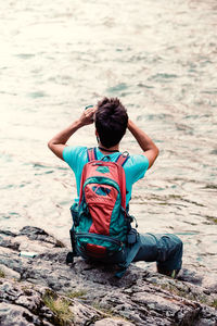 Young tourist with backpack looks through a binoculars, sits on a rock over a river