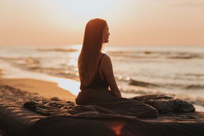 Woman sitting on shore at beach against sky during sunset