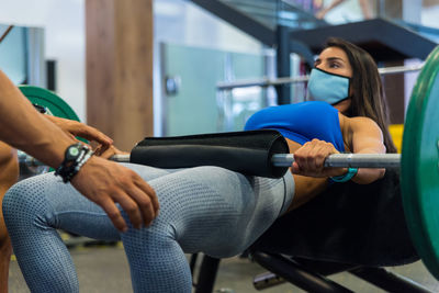 Focused female athlete in mask in sportswear doing hip thrusts with help of professional personal instructor during workout in gym