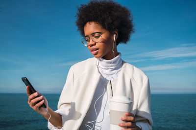 Young woman holding coffee using smart phone against sea