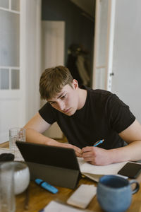 Young man writing while studying from digital tablet at home