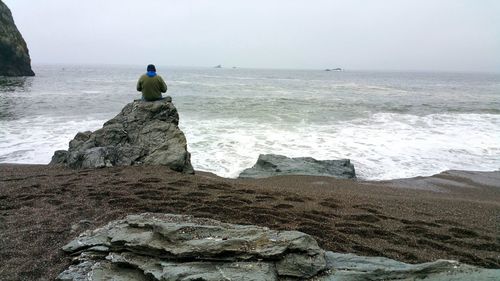 Rear view of man sitting on rock looking at sea against sky