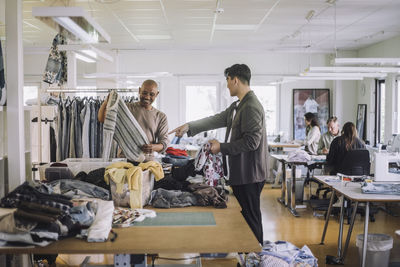 Side view of male fashion designer pointing at colleague holding sweater at workshop