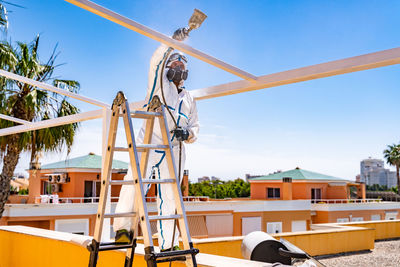 Anonymous employee in protective uniform and mask standing on ladder and using air brush to paint frame structure against cloudy sky on sunny day