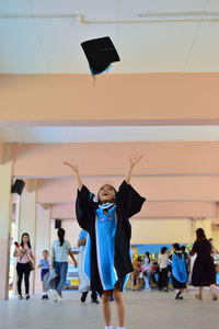 Girl catching mortarboard while standing on floor with people in background