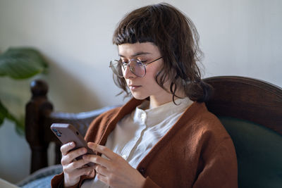 Pensive hipster girl in glasses wearing vintage style looking smartphone screen reading information.