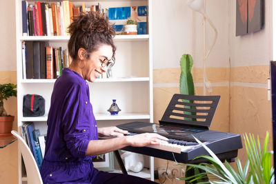 Side view of cheerful young hispanic female playing electric piano while practicing music skills at home