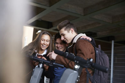 Happy male and female teenage friends with electric push scooters looking at mobile phone below bridge