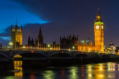 Illuminated bridge over river by buildings against sky at night