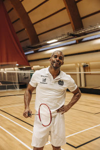 Male badminton player standing with hand on hip while holding racket at sports court