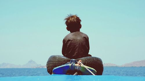 Rear view of boy sitting in boat with tire on sea against clear sky