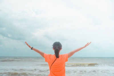 Rear view of woman with arms outstretched standing at beach against sky