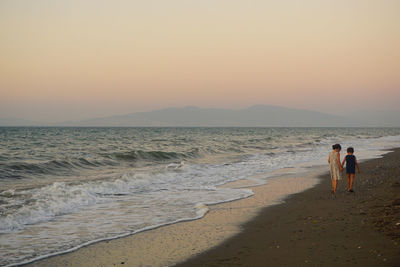 Rear view of siblings walking at beach against clear sky during sunset