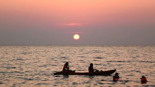 People boating and swimming in sea against sky during sunset
