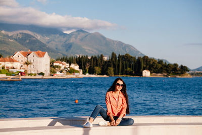 Woman in sunglasses sitting at lake against mountains