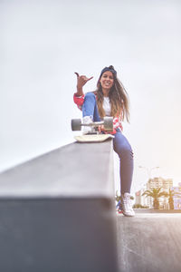 Beautiful young woman in cap is sitting at skatepark on the ramp with her longboard.