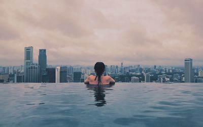 Rear view of woman in infinity pool against cityscape during sunset