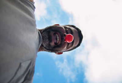 Low angle portrait of man wearing clown nose against cloudy sky