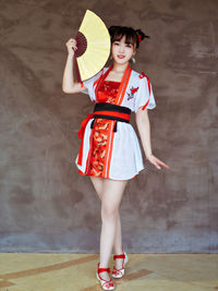 Full length portrait of young woman holding hand fan against wall