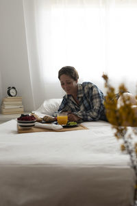 Woman with hair up weared plaid shirt having a breakfast on bed.