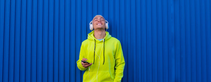 Smiling boy listening music standing by blue wall