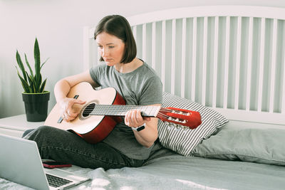 Young man playing guitar while sitting on bed at home